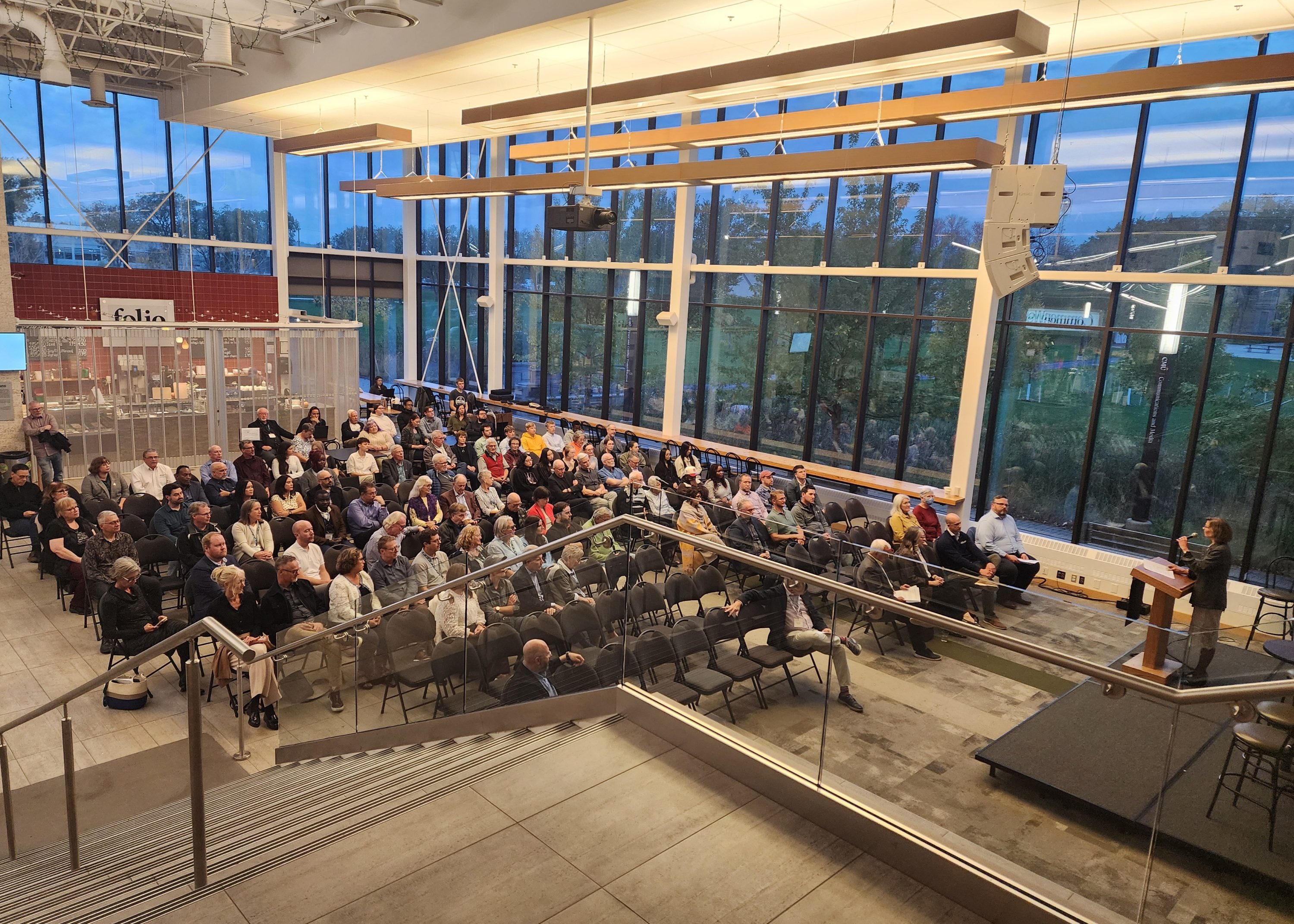Audience seated in a modern lecture hall listening to a speaker at a podium near the front-right corner under glass walls and ceiling lights.